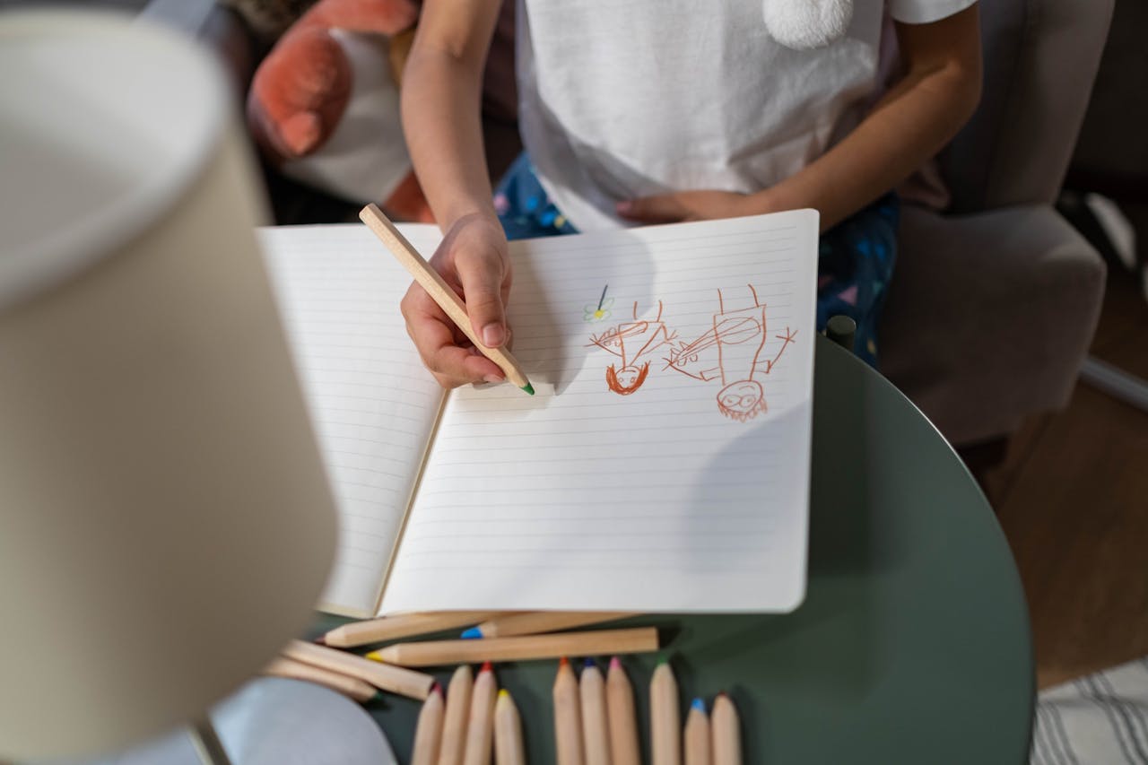 A young child draws in a notebook with colored pencils, creative play indoors.
