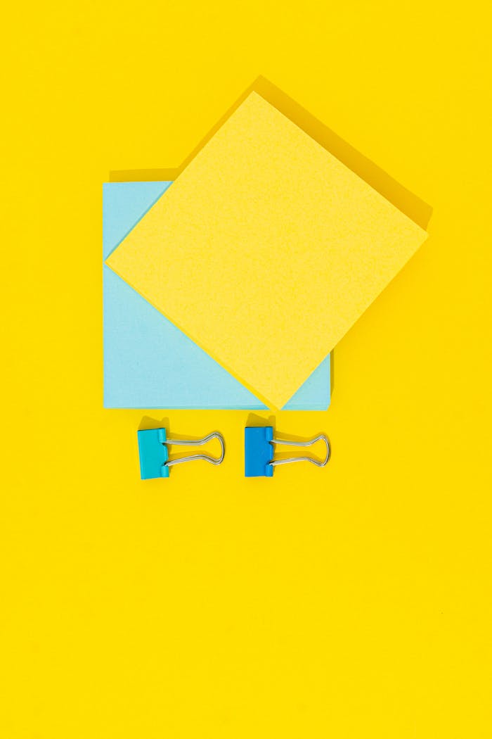 A minimalistic top view of blue binder clips and sticky notes on a bright yellow background.